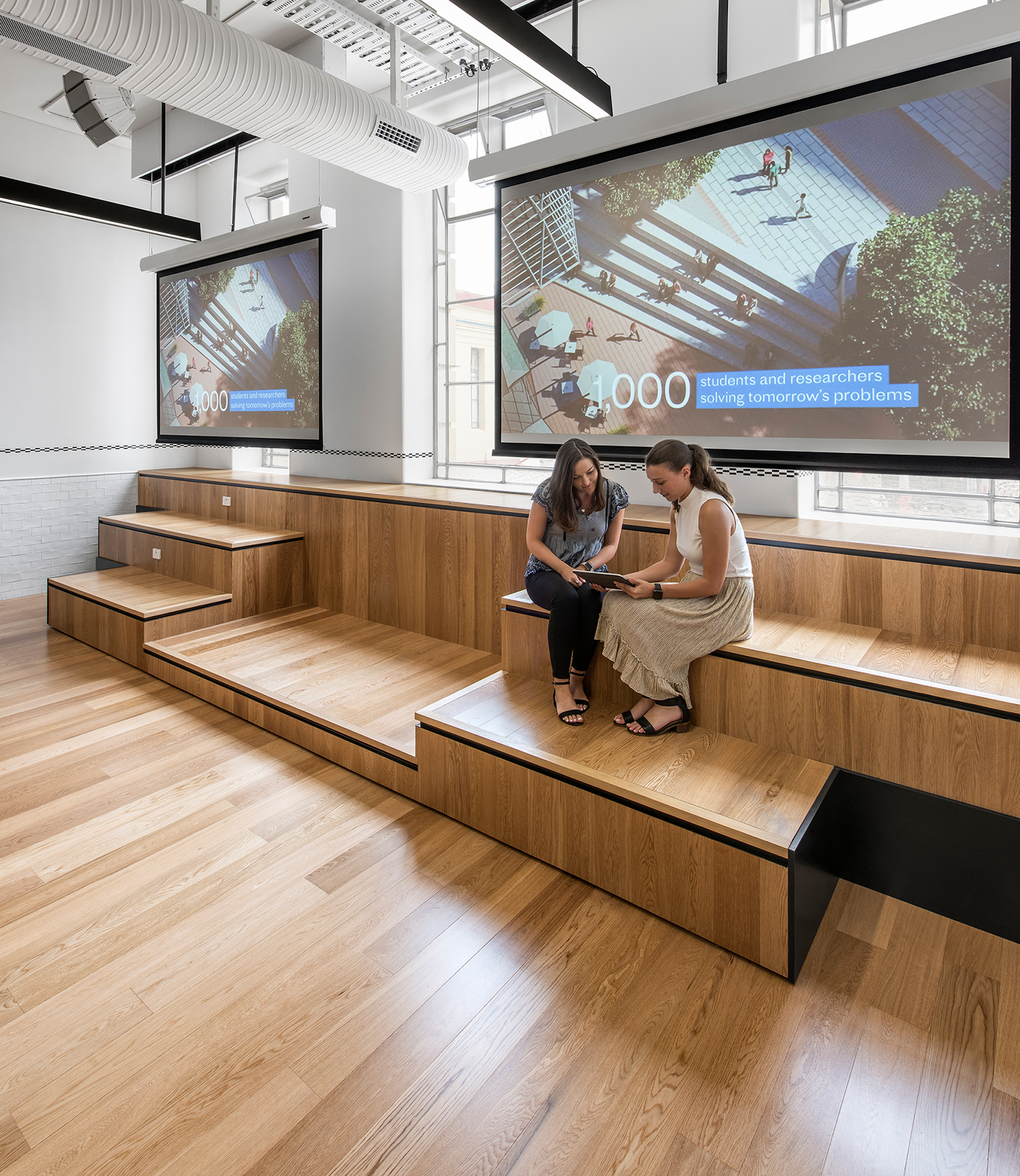 Two people sitting on a wooden bench, two projector screens are behind them