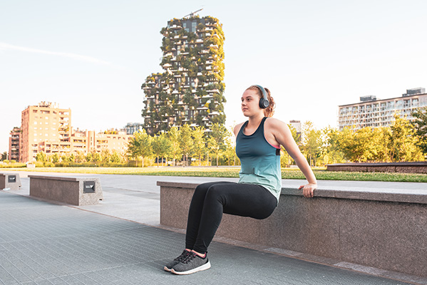 A young woman doing exercise outdoors in a public park, girl of the generation z sports prepares for the summer costume test, copy space, urban skyscrapers background, blu and black sportswear